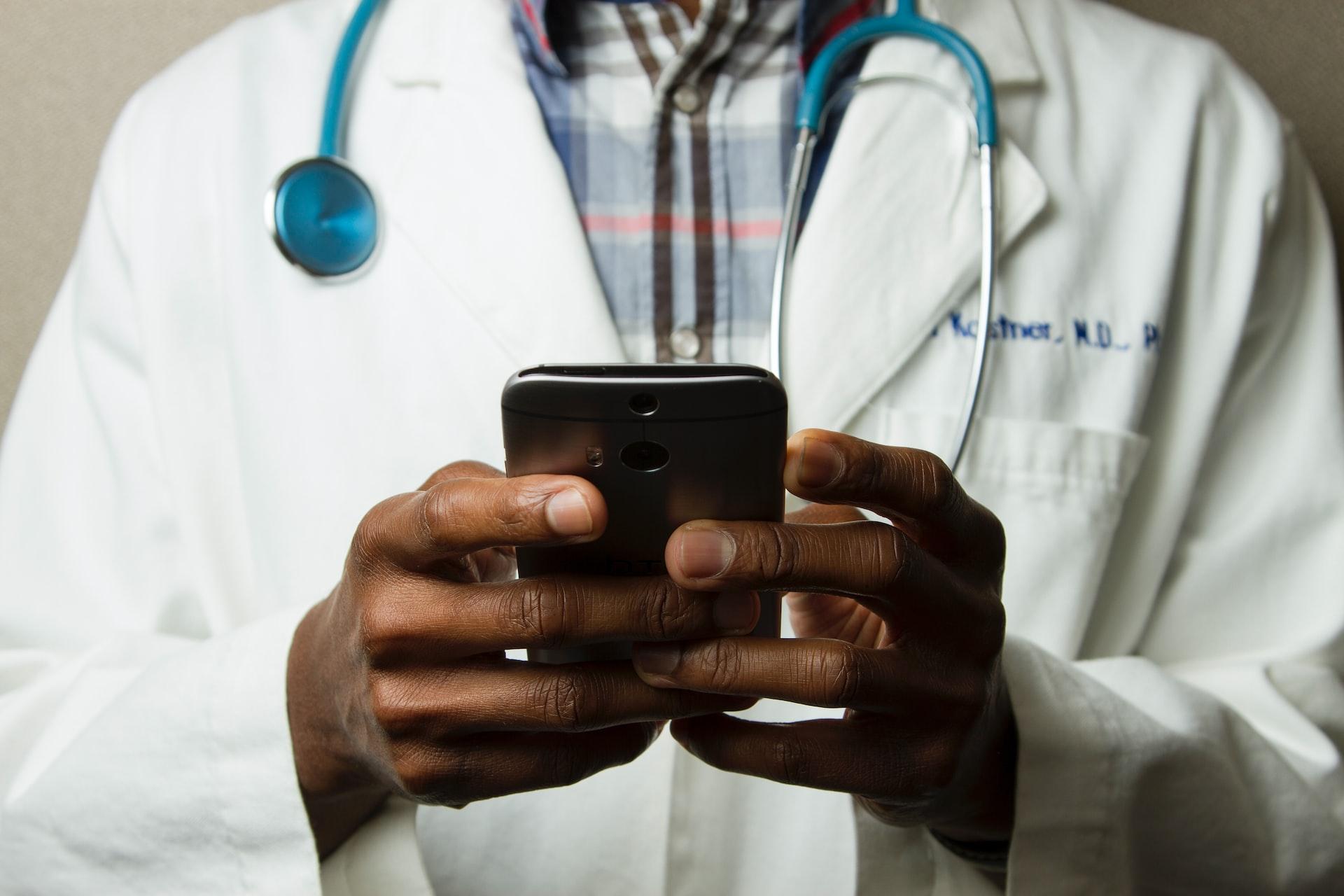 A doctor in a white coat with a stethoscope around their neck is using a smartphone, focusing on the device.